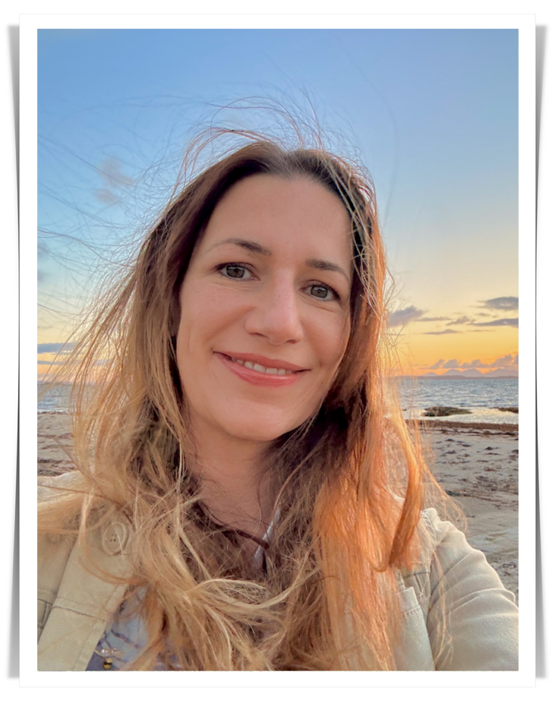 Photograph of a smiling woman with long, windswept hair on a beach, with the sea and sky in the background.