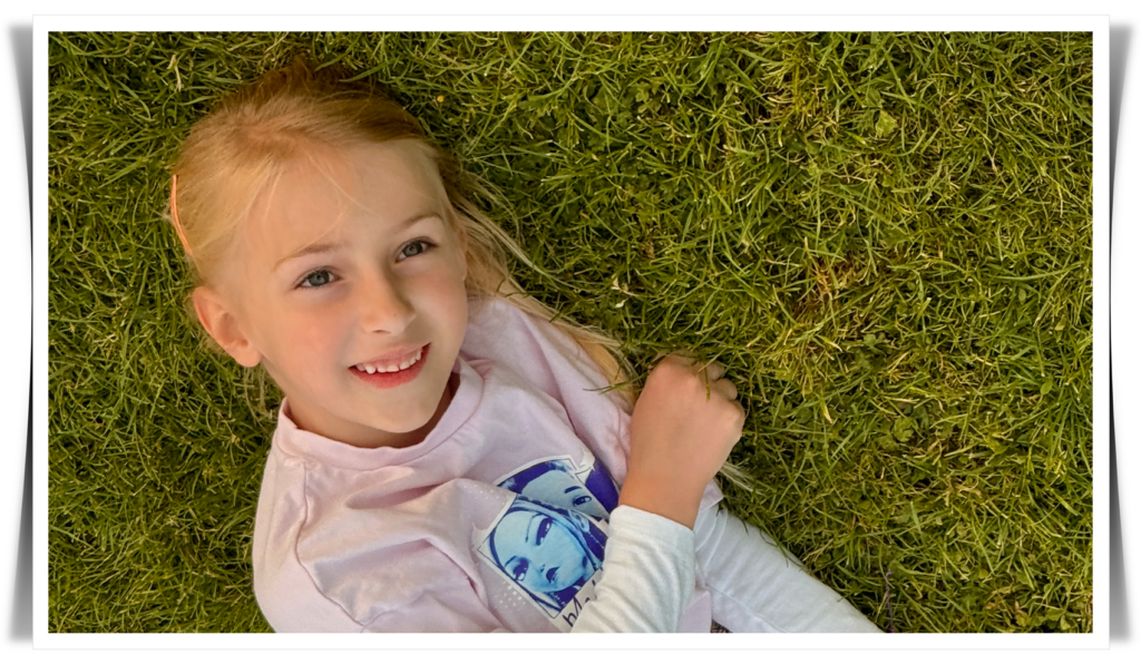 A photograph of a smiling young girl with blonde hair, lying on grass.